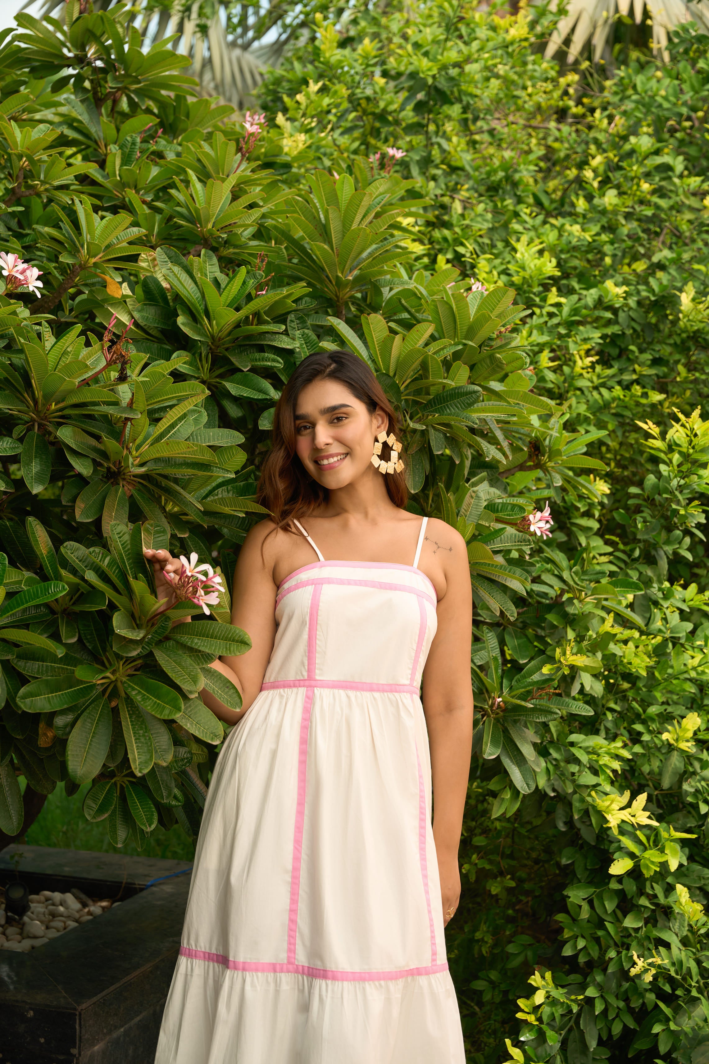Woman wearing a white Amalfi dress with pink trim standing outdoors near green foliage