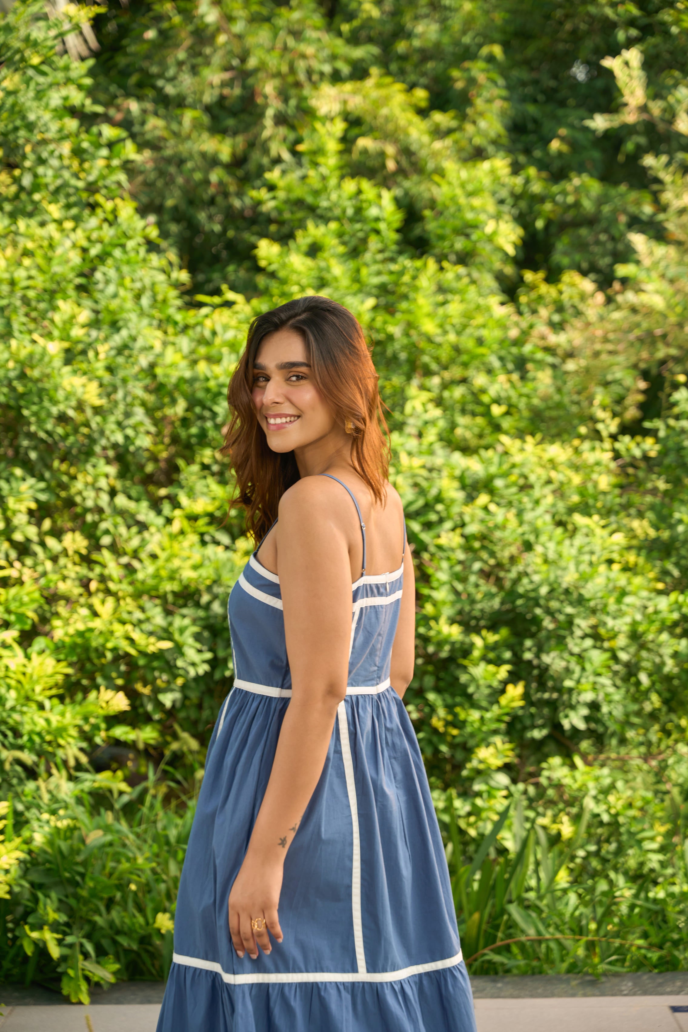 Woman wearing Santorini blue sundress with white trim standing outdoors in front of green foliage
