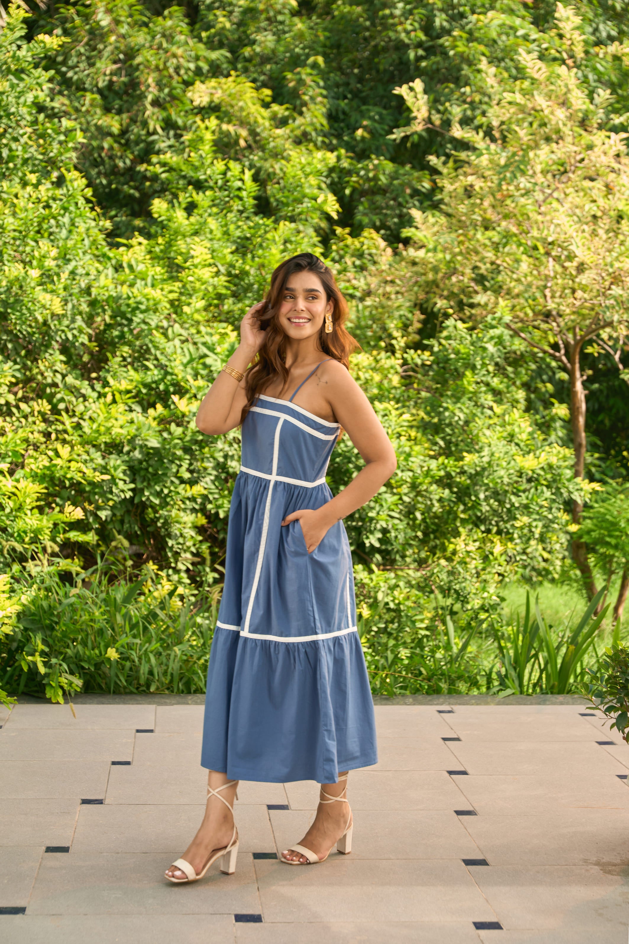 Woman wearing Santorini blue sundress with white trim standing outdoors with greenery background