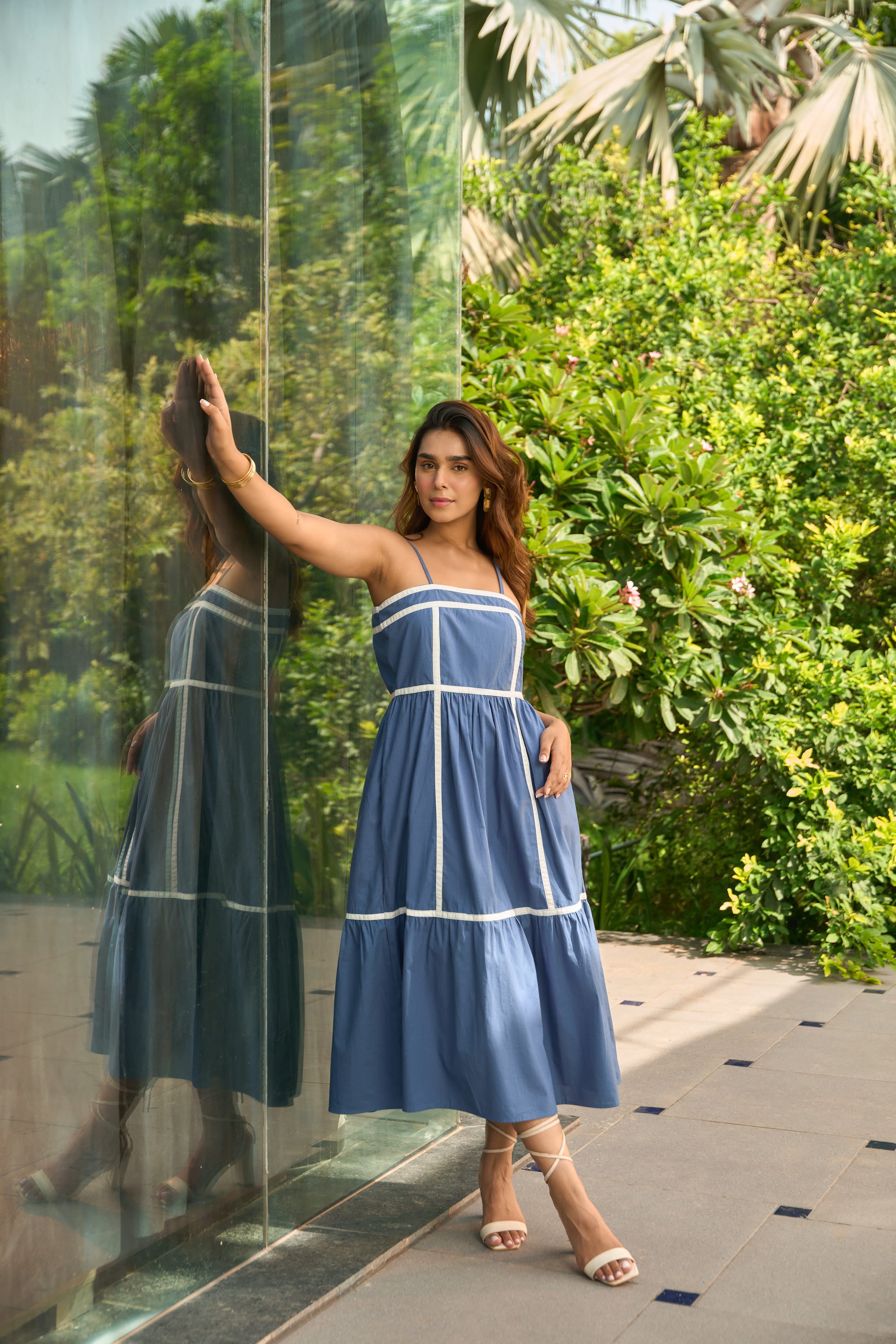 Woman wearing a Santorini blue sundress standing by a glass wall with greenery in the background