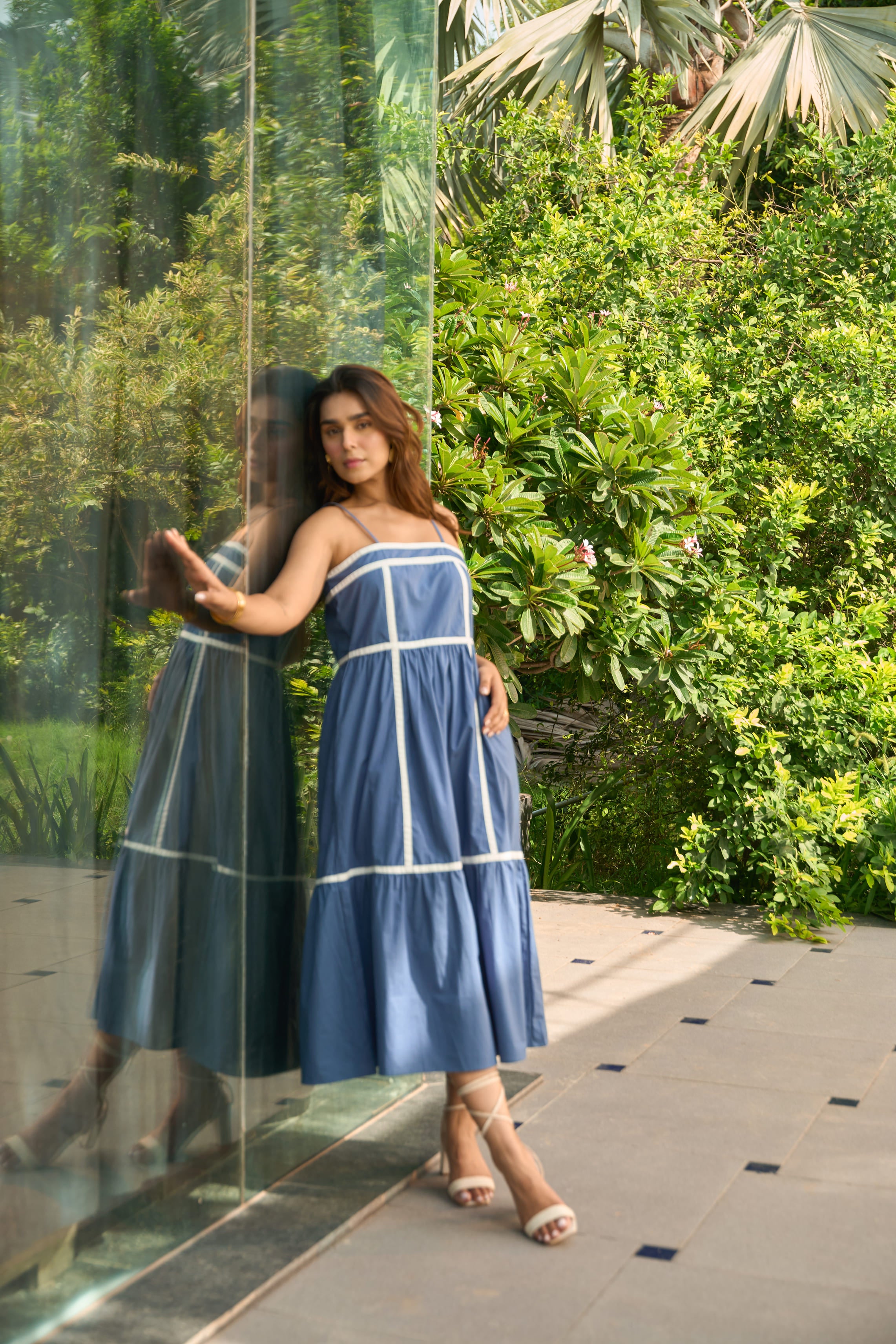 Woman wearing Santorini blue sundress with white trim standing by glass wall outdoors